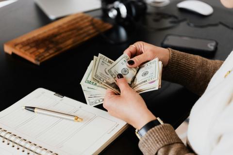 light-skinned hands with black nail polish are holding a stack of $20 bills. the person, body and face out of frame, seem to be sitting at a black desk. there is a calendar planning and pen on the desk.