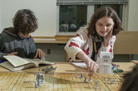 Image depicts two teens playing D&D. One person is looking closely at a D&D rulebook, and one is placing a figurine onto a grid map. 