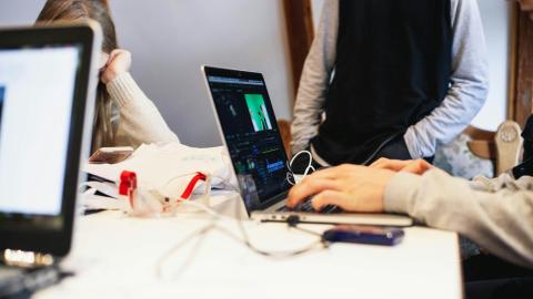 Photo of 3 people sat around a white table, each of them has a computer with editing software open in front of them. There are wires and cords charging the laptops tangled on the table.