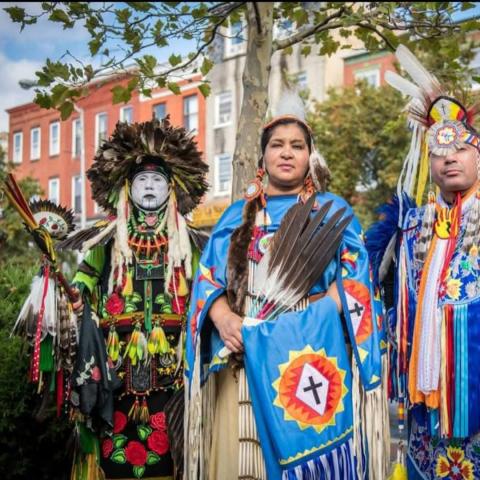 three people in native american traditional clothing stand in a line looking at the camera