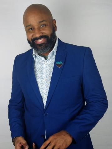 Jonathan McKinney, a black man smiling with a full beard, bald head, wearing a blue blazer and light blue button up shirt.
