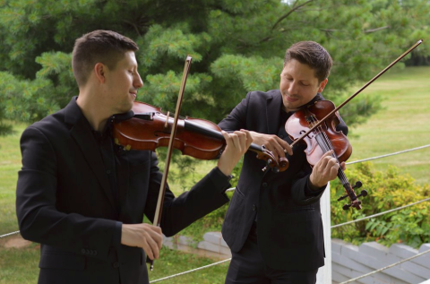 Two young men in black suits playing violin with trees in background
