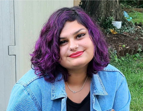 Picture of Alisha, woman smiling with shoulder length curly hair that is purple wearing a jean shirt over a black tshirt.