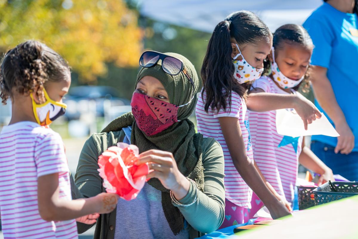 A woman is helping a child with a tissue paper flower craft. Two other children are working on a craft in the background. All are wearing face coverings.