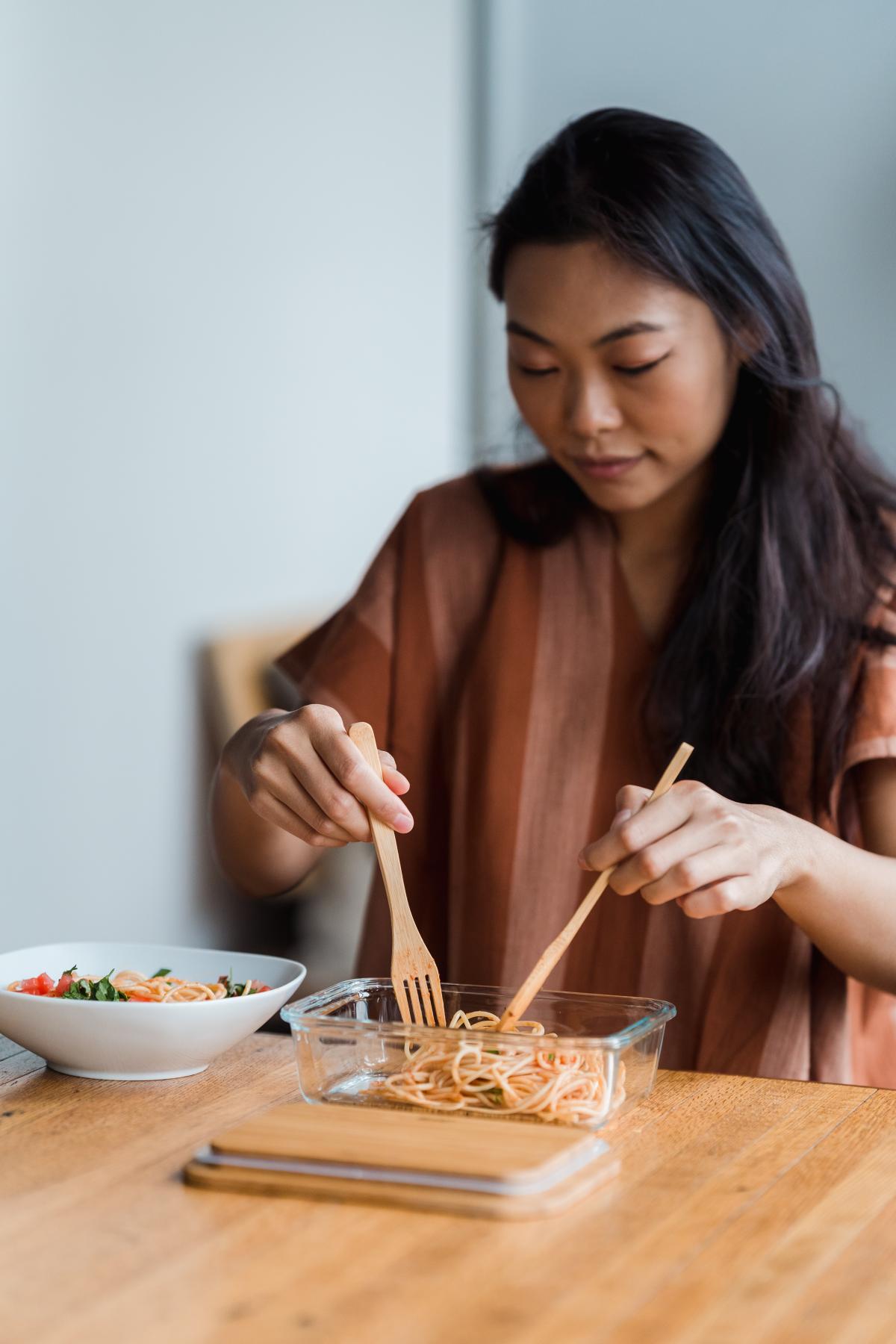 Woman using wooden utensils to move pasta from a plastic storage container to a bowl.