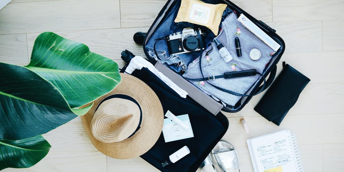 An open suitcase neatly packed next to a palm plant.  Items  included - a camera, sandles and hat. All ready for vacation!