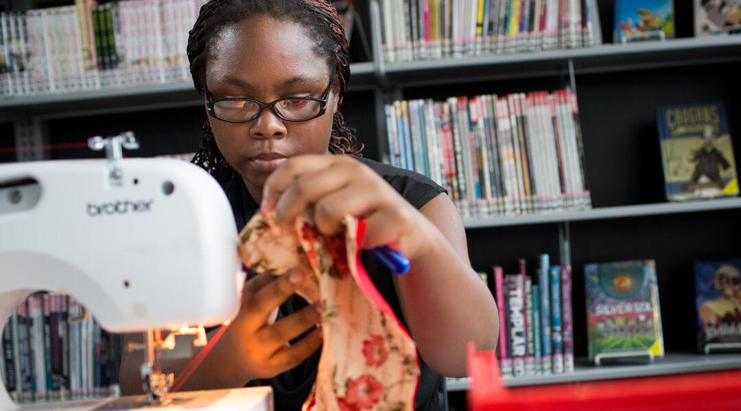 teenager holding fabric next to a sewing machine in front of a background of book shelves