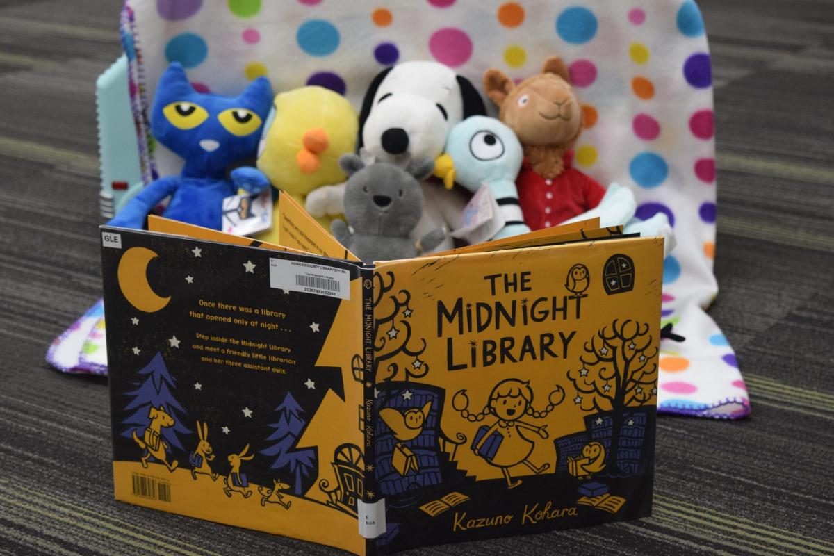 Book characters plush sit on a patterned blanket with "The Midnight Library" open in front of them.