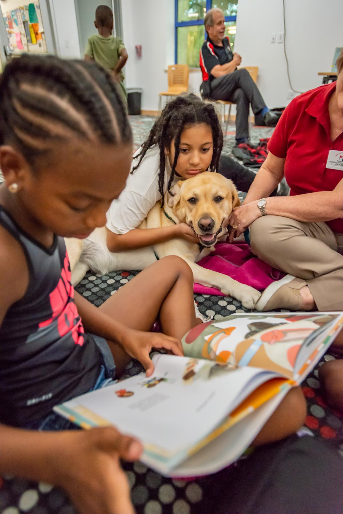 A child hugging a yellow labrador retriever while another child reads a picture book.