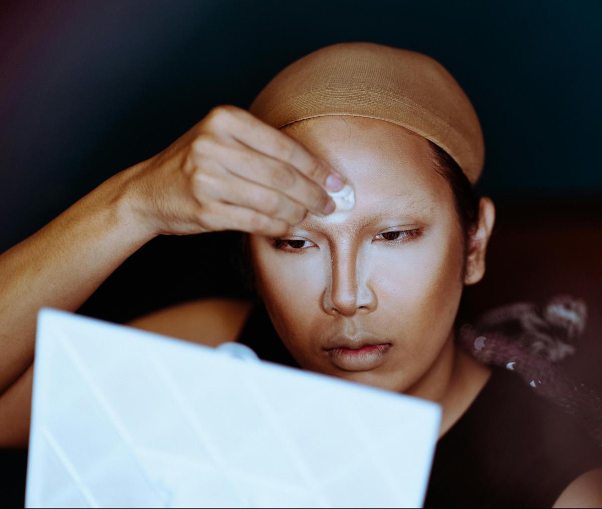 a person of Asian descent, wearing a wig cap, applies face powder to their contoured face.