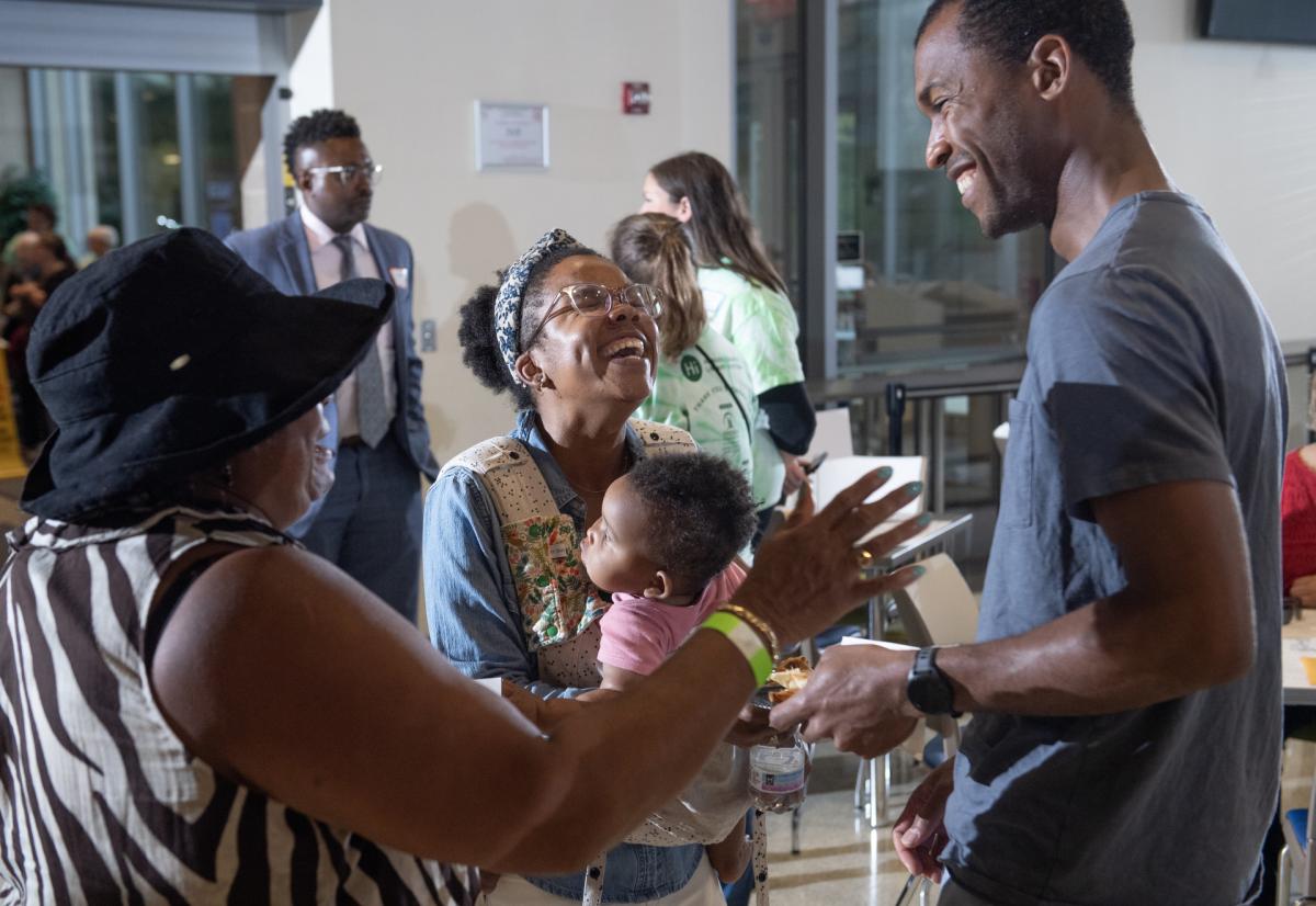 A Black woman holding a baby and laughing, people around her are smiling.