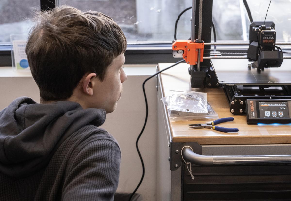a person watches a Prusa MK4S 3D printer that is mid printing a 3D object