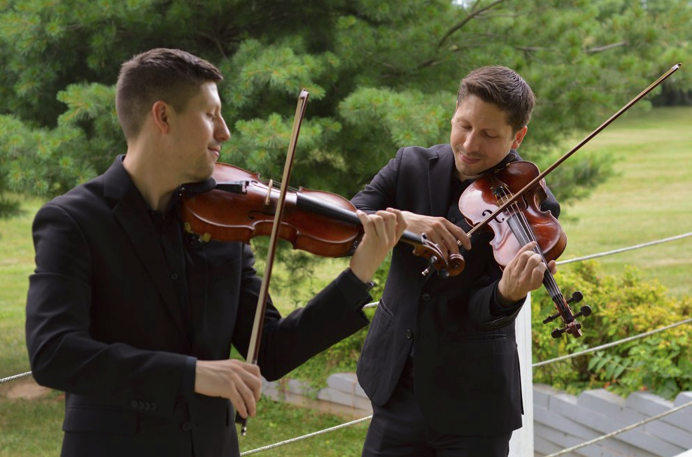 Two young men in black suits playing violin with trees in background