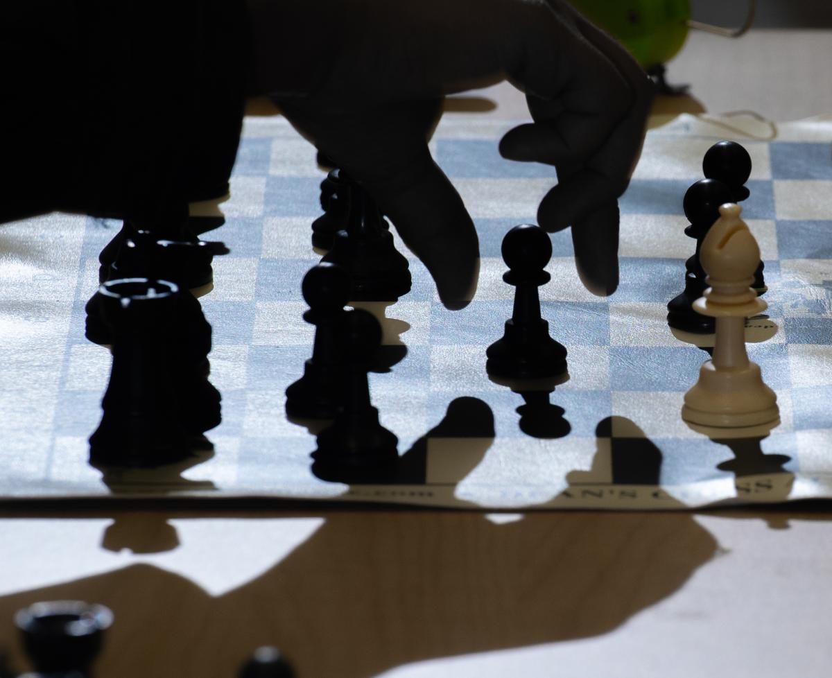 Image depicts a chess board with chess pieces, and a hand reaching to pick up a pawn. 